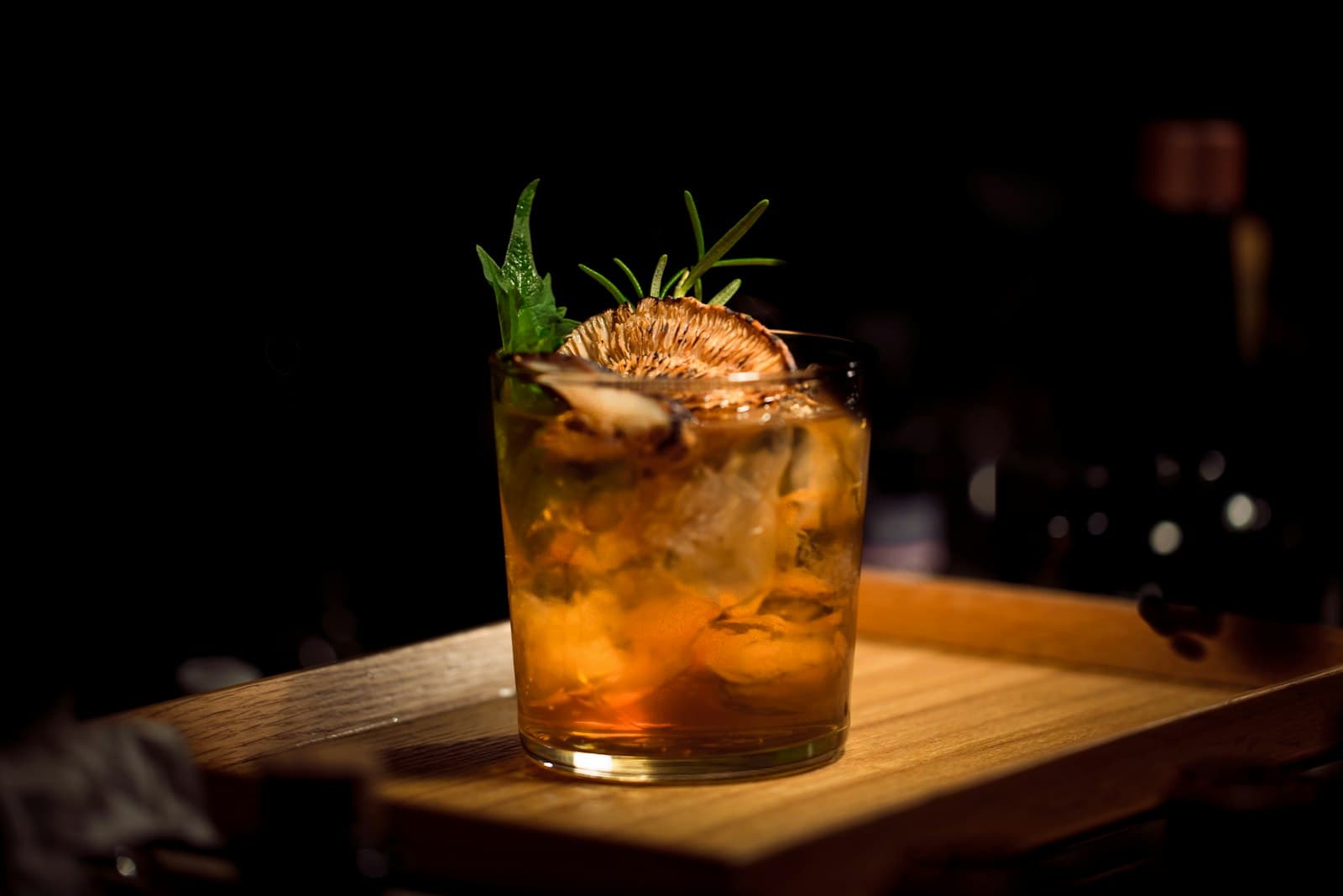 Cocktail ambré au romarin servi sur un plateau en bois dans la pénombre